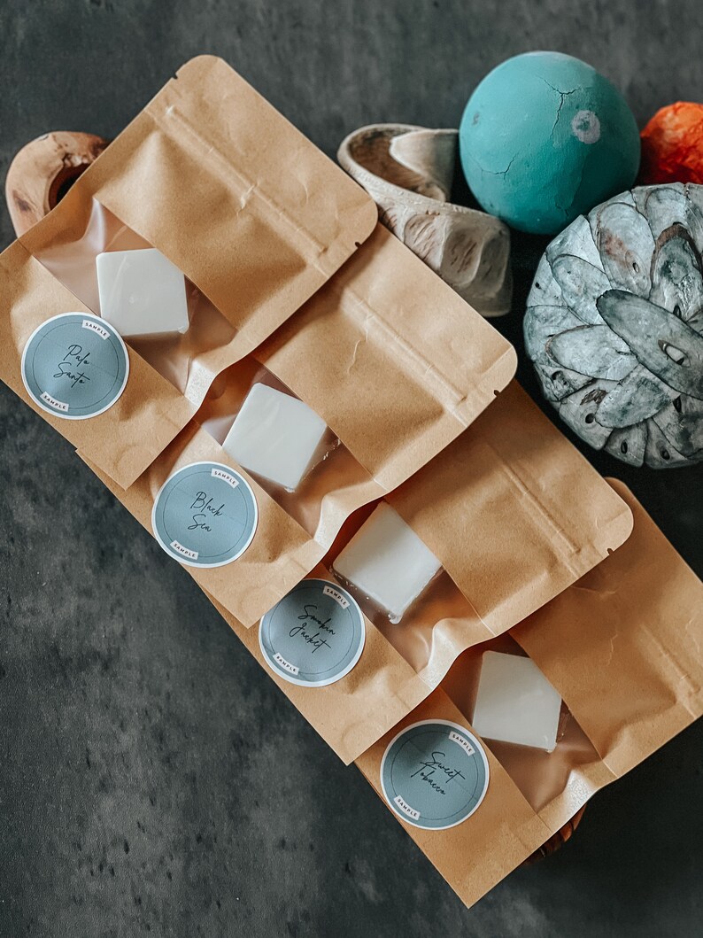 May include: Four brown paper bags containing white wax melts. Each bag features a round sticker with a different scent name: "Pale Sand", "Black Sea", "Smokin' Jacket", and "Sweet Tobacco".