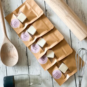 May include: Five small wax melts in individual brown paper bags with clear windows. The wax melts are white and have different scents: Lemon Pound Cake, Oatmeal Cookie, Blueberry Muffin, Almond Brittle, and Granny's Bread Pudding.