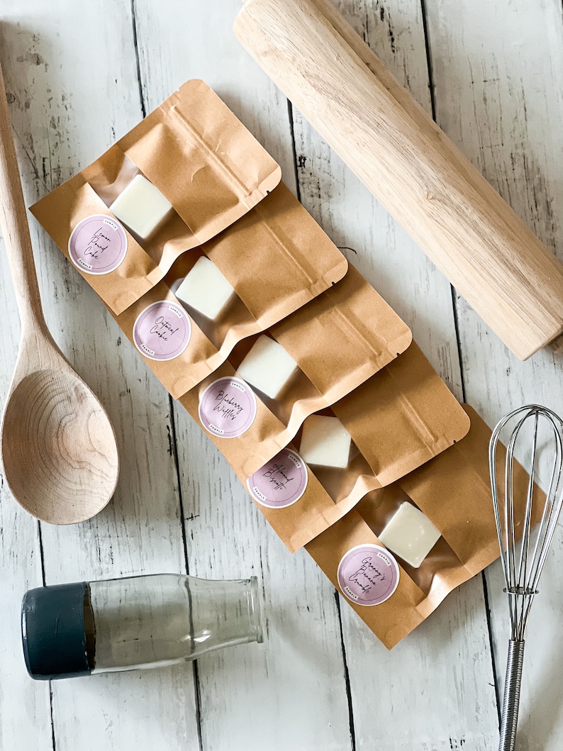 May include: Five small wax melts in individual brown paper bags with clear windows. The wax melts are white and have different scents: Lemon Pound Cake, Oatmeal Cookie, Blueberry Muffin, Almond Brittle, and Granny's Bread Pudding.