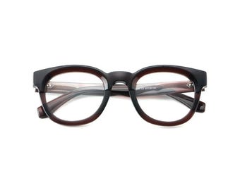 Puede incluir: Gafas con montura marrón oscuro y forma redondeada. Las lentes son transparentes y la montura tiene un diseño de dos tonos, con un tono más oscuro en la parte superior y un marrón más claro en la inferior. Sobre fondo blanco.