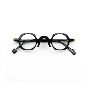 May include: Black eyeglasses with a unique octagonal frame design. The glasses have clear lenses, black arms, and small yellow accents on the temples. The overall style is modern and distinctive.