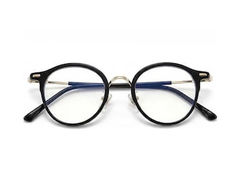 Pode incluir: Óculos de armação redonda com armação preta e detalhes dourados. As lentes são transparentes e as hastes são pretas. Os óculos têm um design clássico e minimalista.