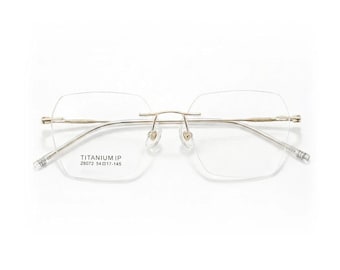May include: Rimless eyeglasses with a gold-tone bridge and temples. The lenses are a geometric shape. The text "TITANIUM IP Z6072 54 17-145" is printed on the left lens.