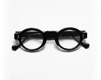 May include: Black round-frame eyeglasses with a keyhole bridge and small, rounded temples. The glasses have a minimalist design and are set against a white background. The lenses are clear, and the frames are a solid, matte black.