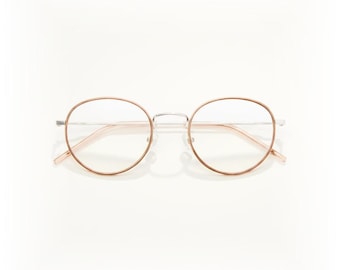 Puede incluir: Gafas redondas con una montura fina en oro rosa y lentes transparentes. El puente y las patillas son plateados. Las gafas están sobre un fondo blanco.