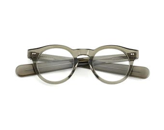 Könnte beinhalten: Graue Brille mit runden Gläsern, Schlüssellochsteg und dicken Bügeln. Die Brille besteht aus einem durchscheinenden Material, und die Gläser sind klar. Der Rahmen ist etwa 14 cm breit.