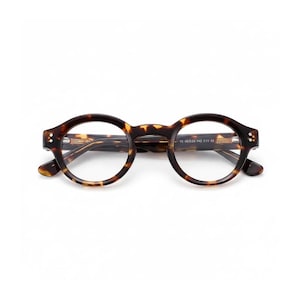 Puede incluir: Gafas de montura redonda con un patrón de carey. Las gafas tienen un diseño clásico con una combinación de colores marrón oscuro y ámbar. La montura presenta pequeños remaches en las patillas y el texto "19 42020-142 C11 CE".