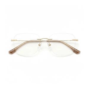 May include: Rimless eyeglasses with a gold-colored metal frame and clear lenses. The temples and nose bridge are a light brown color. The glasses have a square shape and are set against a white background.