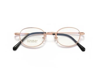 May include: Rose gold oval eyeglasses with black arms. The glasses have a thin frame and the text "TITANIUM SP446 4022 140L" is printed on the left lens. The lenses are clear.