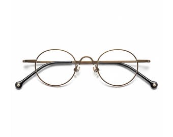 Op de afbeelding: Ronde bril met een bronzen montuur en zwarte pootjes. De bril heeft een klassiek, minimalistisch ontwerp met heldere glazen. De pootjes hebben een klein, decoratief detail aan het uiteinde.