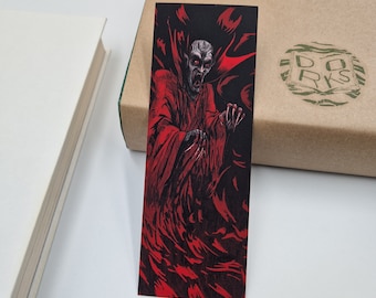 Könnte beinhalten: Schwarzer Lesezeichen mit einer rot-schwarzen Illustration eines Vampirs. Der Vampir wird mit einem blassen Gesicht, roten Augen und einem roten Gewand dargestellt. Das Lesezeichen ist rechteckig und befindet sich neben einem Buch und einer Pappschachtel.