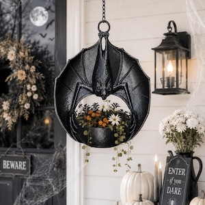 May include: A black hanging planter shaped like a bat with its wings spread wide, holding a circular bowl containing small orange and white flowers with trailing green vines. The bat is suspended by a metal chain from a circular top ring.