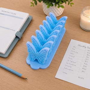 May include: A light blue desk organizer featuring a series of five connected butterfly-shaped slots. The organizer is made of a solid material and is designed to hold papers or cards in an upright position, arranged in a linear row on a flat base.