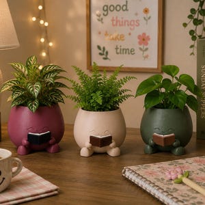 May include: Three rounded ceramic planters in burgundy, light pink, and dark green, each shaped like a seated figure holding a small book. Each planter contains a green plant, with the figures featuring simple carved faces and small feet.