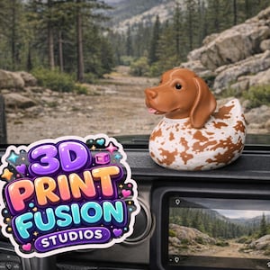 May include: A brown and white spotted dog-shaped rubber ducky sits on a car dashboard. The duck has brown ears and a pink tongue. The dashboard also displays a colorful sticker with the text "3D PRINT FUSION STUDIOS". The background shows a scenic mountain road.