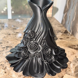 May include: A matte black vase shaped like a flared dress with a cinched waist and a wide, ruffled hem. The surface is decorated with multiple three-dimensional sculpted roses of varying sizes that wrap around the lower portion of the object.