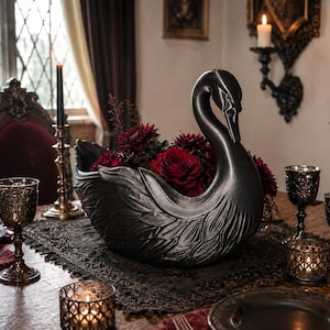 May include: A matte black swan-shaped bowl featuring detailed sculpted feathers on its exterior. The bowl is filled with deep red flowers and is positioned as a centerpiece on a table, showcasing a curved neck and a hollow interior for floral arrangements.