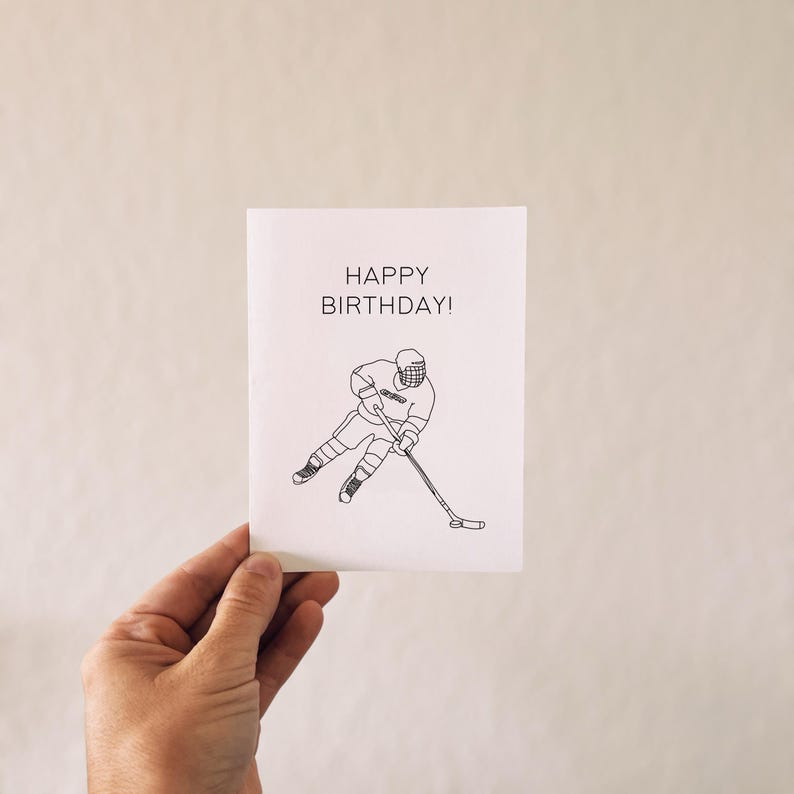 May include: A white card with black line art of a hockey player skating and holding a stick. The text "HAPPY BIRTHDAY!" is printed in black at the top of the card.
