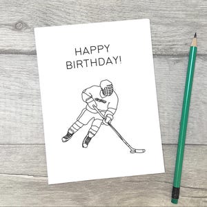 May include: A black and white illustration of a hockey player on a white card with the text "Happy Birthday!" printed on it.