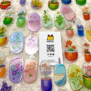 Puede incluir: Surtido de pegatinas transparentes en forma de cápsula de colores, con diferentes diseños florales. Cada pegatina tiene una flor y una planta diferentes en su interior, sobre un fondo blanco. Las pegatinas están dispuestas sobre una superficie blanca con una tarjeta de visita para "puddingcat", papelería y regalos.