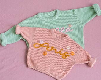 May include: Two knitted baby sweaters, one pink with the name "Aris" embroidered in yellow with small white flowers, and one light green with the name "Lea" embroidered in pink with a small green flower.