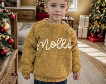 May include: A young boy wearing a yellow knitted sweater with the name "mollie" embroidered in white. He is standing in front of a Christmas tree.
