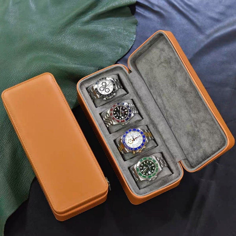 Op de afbeelding: Een bruine leren horlogebox met vier vakken voor horloges. De doos is open en vier horloges zijn zichtbaar. De horloges hebben zilveren banden en zwarte wijzerplaten met witte cijfers en wijzers. De horloges hebben verschillende gekleurde accenten rond de wijzerplaat.