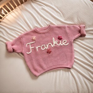 May include: A pink knitted jumper with the name "Frankie" embroidered on the front in white yarn. The jumper has small flower embellishments in pink and white.