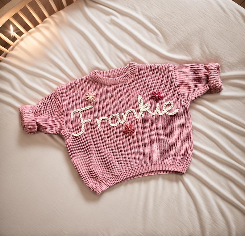 May include: A pink knitted jumper with the name "Frankie" embroidered on the front in white yarn. The jumper has small flower embellishments in pink and white.