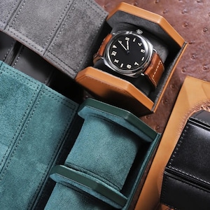 May include: A luxury watch with a black face and brown leather band rests in a brown leather watch case. The watch case is open, revealing a gray interior. Other watch cases in gray, green, and black leather are also visible.