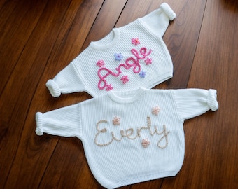 May include: Two white knitted baby sweaters with embroidered names. The top sweater has the name "Angle" embroidered in pink and purple thread. The bottom sweater has the name "Everly" embroidered in gold thread.