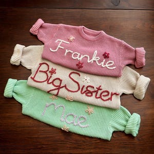 May include: Three knitted jumpers stacked on top of each other. The top jumper is pink with the name "Frankie" embroidered in white. The middle jumper is beige with the words "Big Sister" embroidered in red. The bottom jumper is green with the word "Mae" embroidered in purple.
