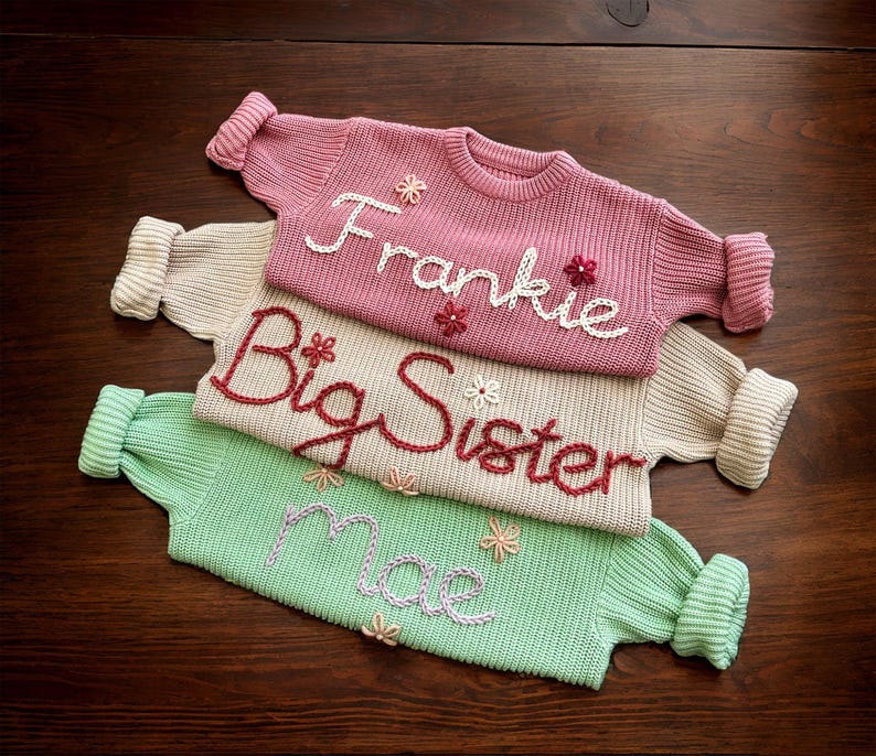 May include: Three knitted jumpers stacked on top of each other. The top jumper is pink with the name "Frankie" embroidered in white. The middle jumper is beige with the words "Big Sister" embroidered in red. The bottom jumper is green with the word "Mae" embroidered in purple.