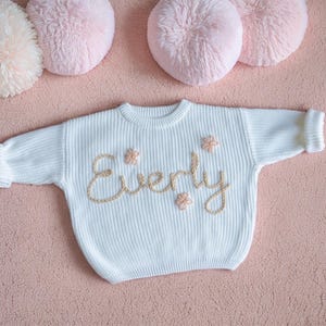 May include: White knitted sweater with the name "Everly" embroidered in gold thread and small pink flowers.