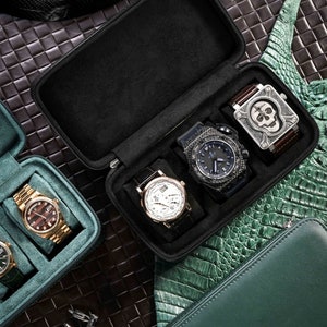 Op de afbeelding: Een zwarte horlogekoffer met vier horloges erin. De horloges hebben verschillende stijlen en kleuren, waaronder een zilveren horloge met een witte wijzerplaat, een zwart horloge met een blauwe wijzerplaat en een zilveren horloge met een zwarte wijzerplaat. De koffer is open en de horloges zijn zichtbaar.