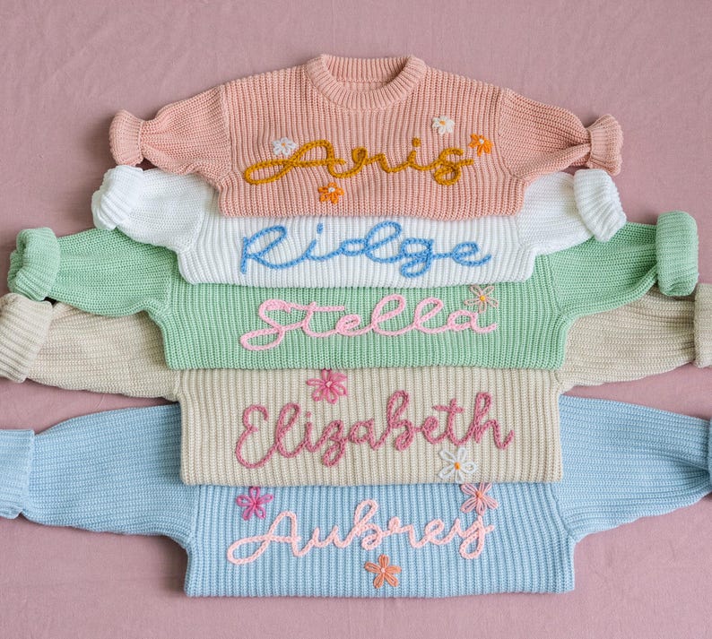 May include: A stack of five knitted jumpers in different pastel colours, each with a different name embroidered on the front in a cursive font. The names are: Aris, Ridge, Stella, Elizabeth, and Aubrey.
