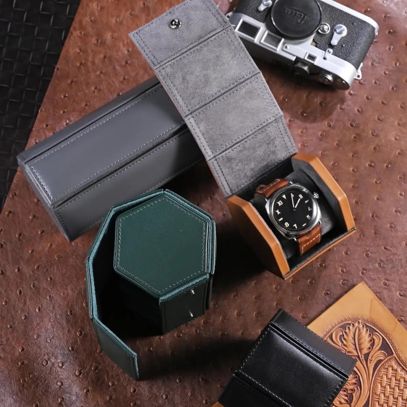 May include: An assortment of leather watch cases in various colors, including gray, green, and black. One case holds a brown leather-strapped watch with a black face. A vintage camera is visible in the background.