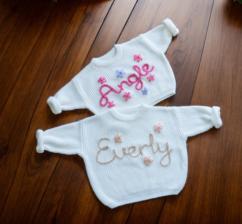 May include: Two white knitted baby sweaters with embroidered names. The top sweater has the name "Angle" embroidered in pink and purple thread. The bottom sweater has the name "Everly" embroidered in gold thread.
