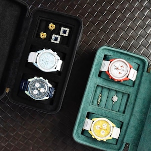 Op de afbeelding: Drie horlogedozen met verschillende horloges en sieraden. De dozen zijn gevoerd met groen vilt en hebben een zwarte buitenkant. De horloges hebben witte, rode en gele wijzerplaten.