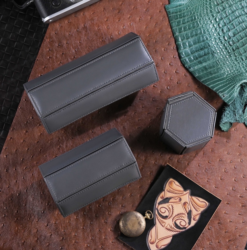 May include: Three gray leather watch cases and a hexagonal watch holder are displayed on a brown textured surface. A vintage pocket watch and a leather-tooled design are also present. The cases are rectangular with a hinged design.
