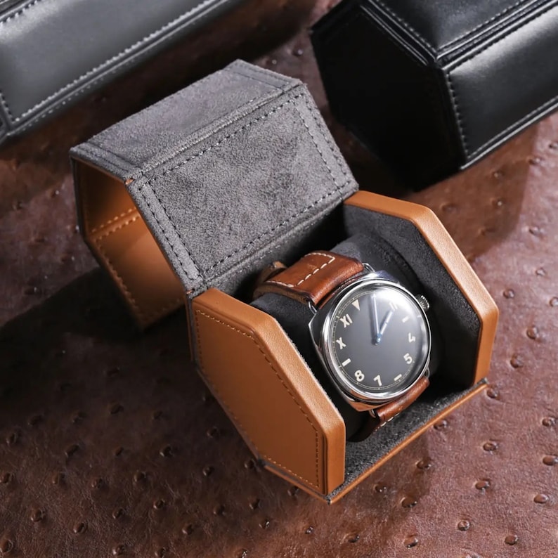 May include: A brown leather watch with a black face and a brown leather strap rests inside an open, hexagonal watch case. The case is gray suede on the inside and brown leather on the outside. Additional black watch cases are visible in the background.