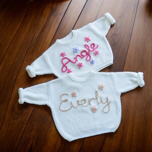 May include: Two white knitted baby sweaters with embroidered names. The top sweater has the name "Angle" embroidered in pink and purple thread. The bottom sweater has the name "Everly" embroidered in gold thread.