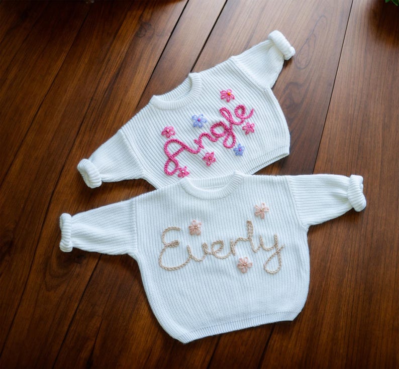 May include: Two white knitted baby sweaters with embroidered names. The top sweater has the name "Angle" embroidered in pink and purple thread. The bottom sweater has the name "Everly" embroidered in gold thread.