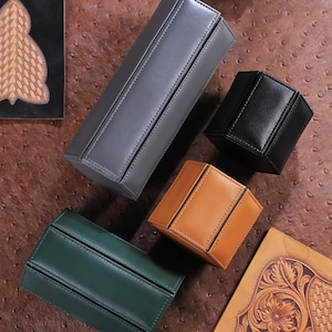May include: An assortment of leather boxes in various colors: gray, black, tan, and green. The boxes have a geometric design with stitched seams. The boxes are arranged on a textured brown surface.
