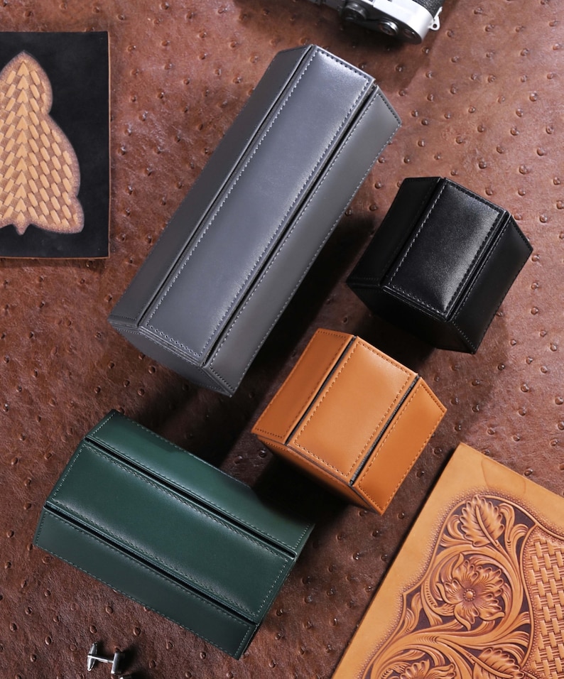 May include: An assortment of leather boxes in various colors: gray, black, tan, and green. The boxes have a geometric design with stitched seams. The boxes are arranged on a textured brown surface.