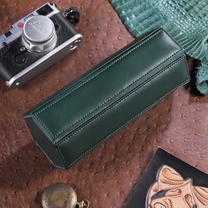 May include: A dark green leather rectangular case with white stitching. A vintage camera, a pocket watch, and a leather piece are on a brown textured surface.