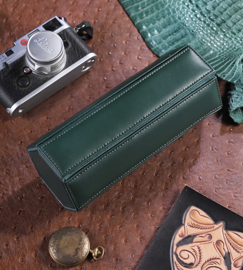 May include: A dark green leather rectangular case with white stitching. A vintage camera, a pocket watch, and a leather piece are on a brown textured surface.