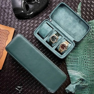 Op de afbeelding: Twee groene leren horlogedozen, een met een scharnierend deksel en drie vakken voor horloges, de andere een lange, rechthoekige doos. De doos met het deksel heeft twee horloges erin, een met een gouden wijzerplaat en een met een zwarte wijzerplaat.