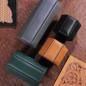 May include: An assortment of leather boxes in various shapes and colors, including gray, black, tan, and green. The boxes are arranged on a textured brown surface, with visible stitching details and a minimalist design. The boxes appear to be for storage or display.