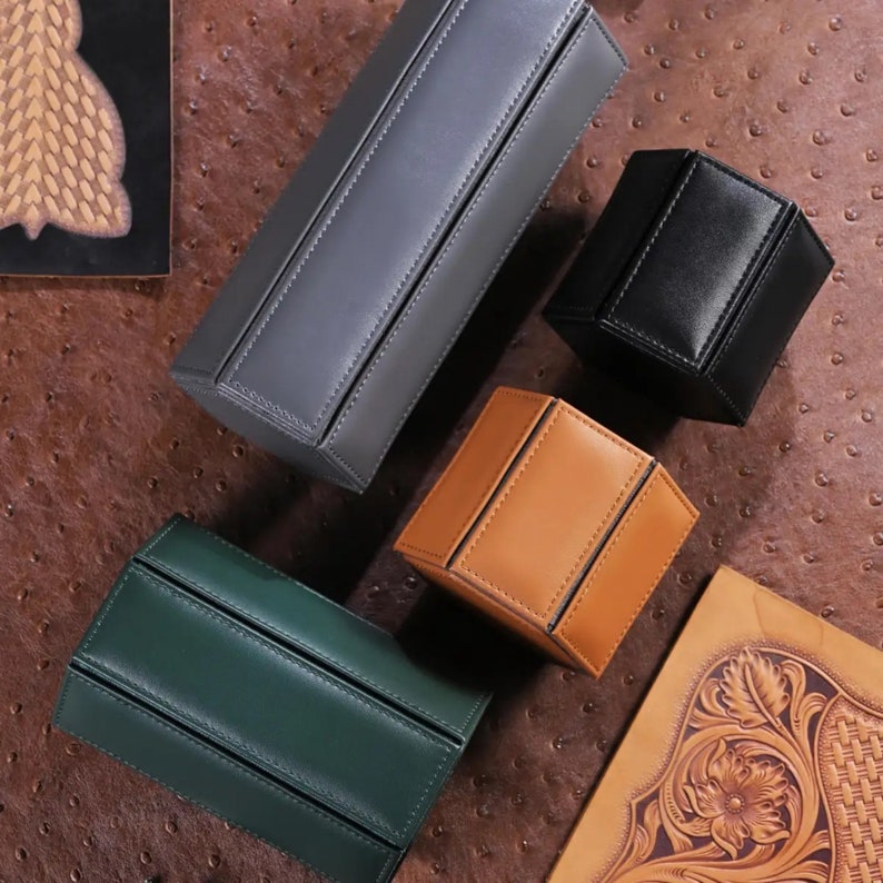 May include: An assortment of leather boxes in various shapes and colors, including gray, black, tan, and green. The boxes are arranged on a textured brown surface, with visible stitching details and a minimalist design. The boxes appear to be for storage or display.